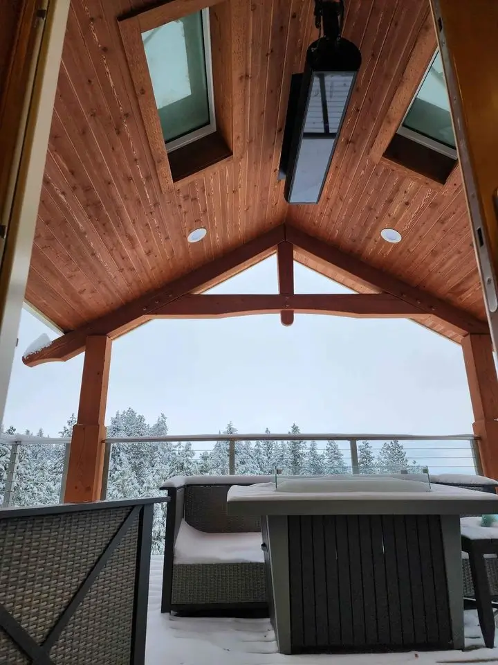 A snowy outdoor deck with a wooden roof, covering with skylights, and a large heater overhead. There are wicker sofas and a table, with a scenic view of snow-covered trees in the background.