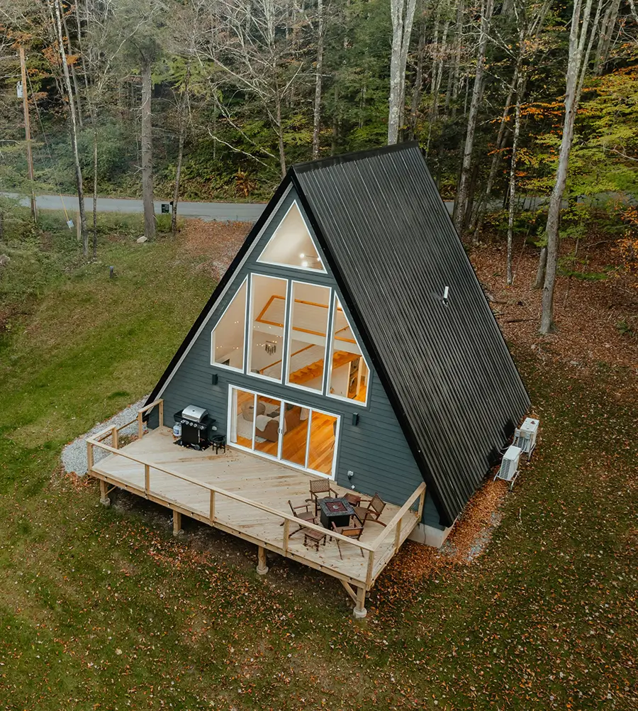 A-frame cabin with large glass windows and a wooden deck, surrounded by grass and autumn trees, nestled in a quiet, wooded area.