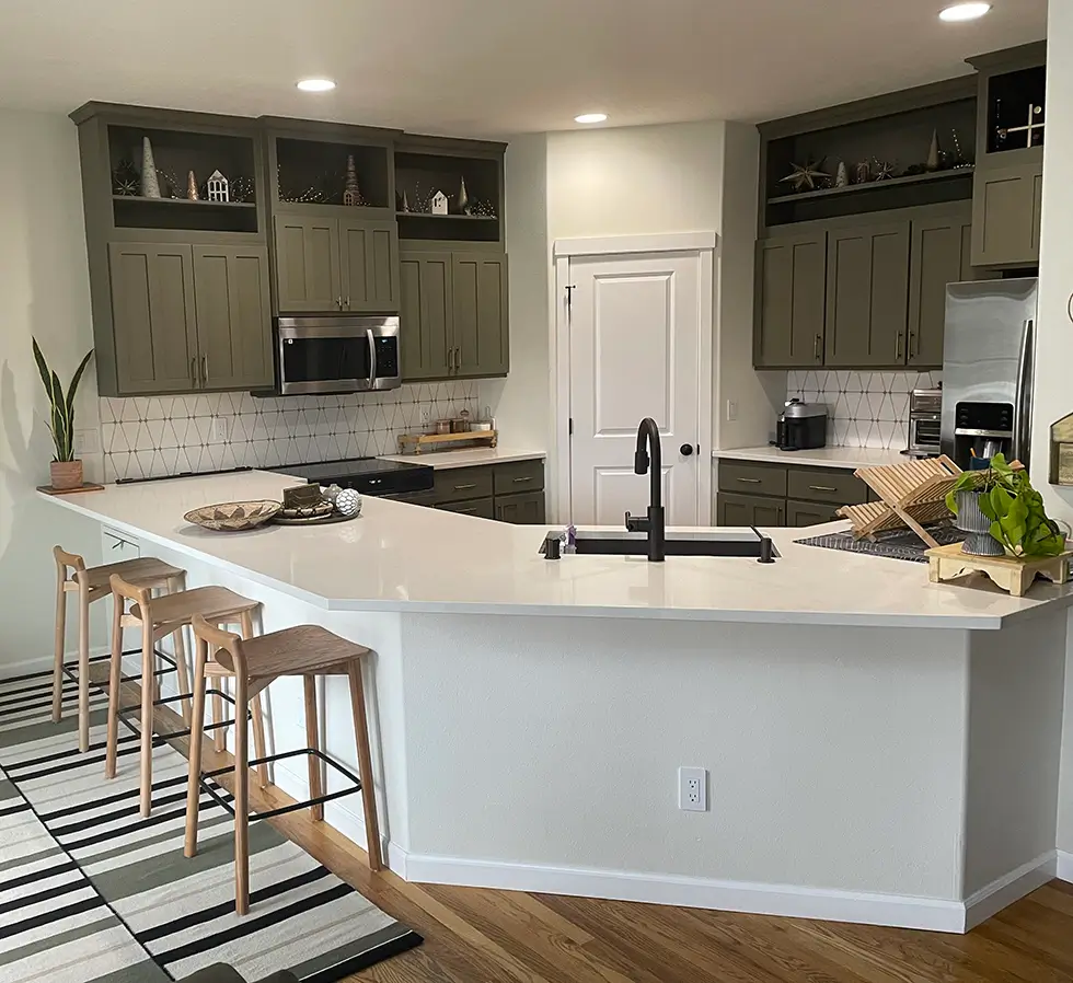 Modern kitchen with olive green cabinets, white countertops, and a geometric backsplash. Three wooden bar stools line the island, and open shelves above display decor. A striped rug covers part of the wooden floor.