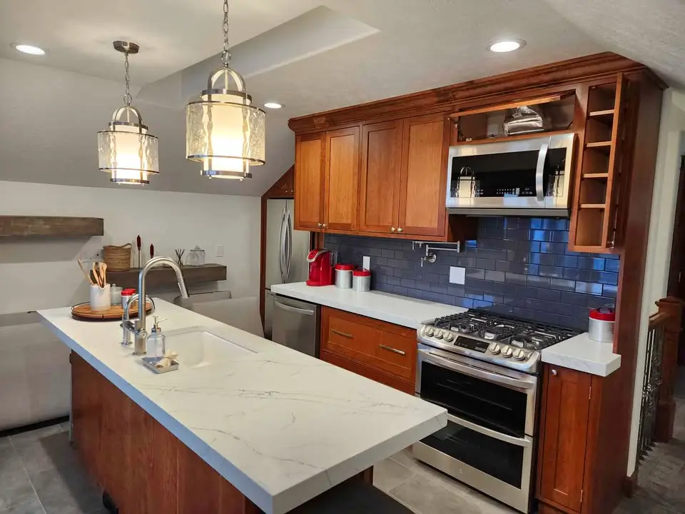 Modern kitchen with wood cabinets, stainless steel appliances, a marble island with a sink, hanging lights, and a bluish gray tile backsplash.